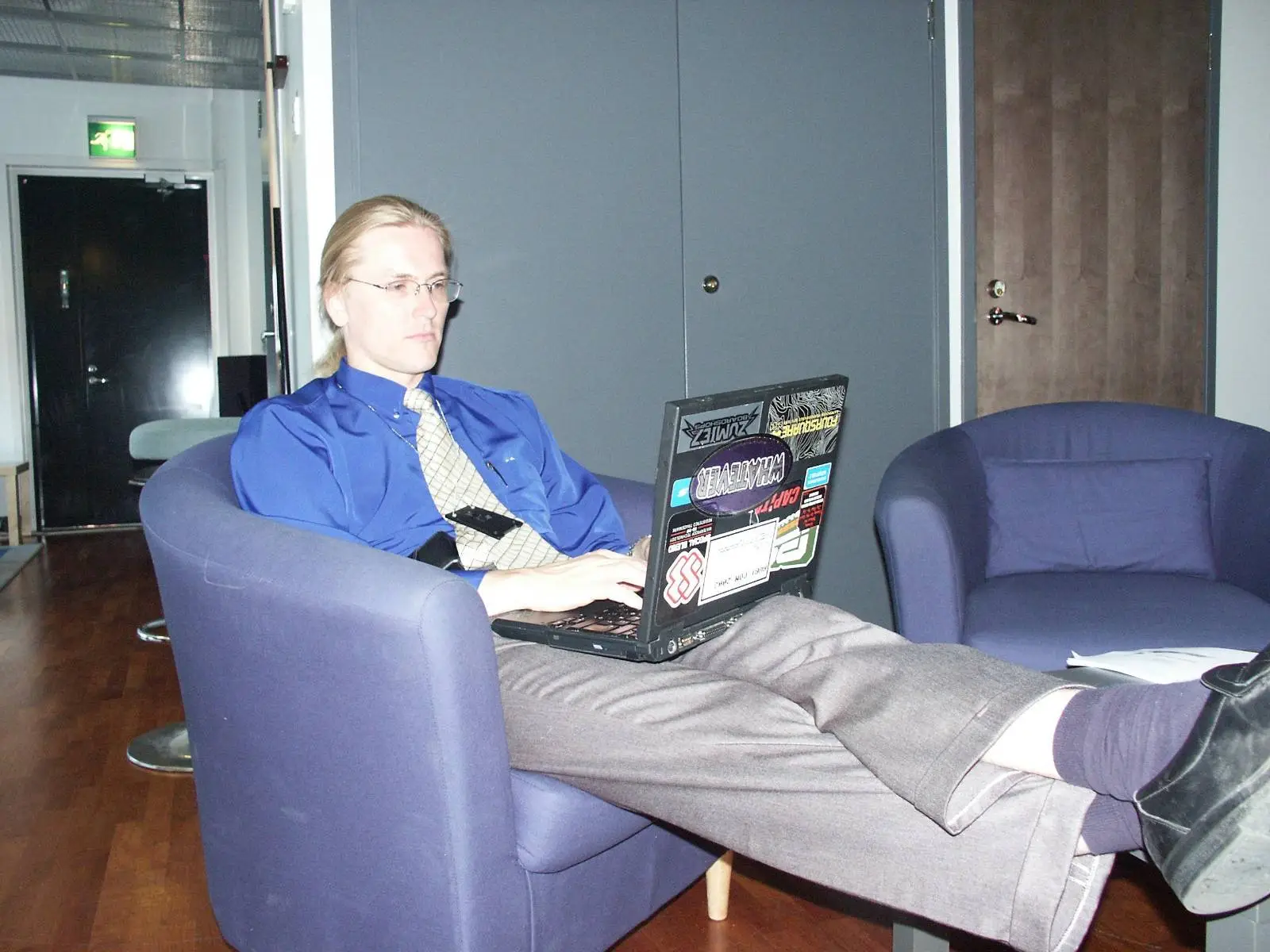 a photo of young Mikko Hyppönen, wearing a blue shirt and tie, on a purple chair, his feet up and a laptop with stickers on his lap.